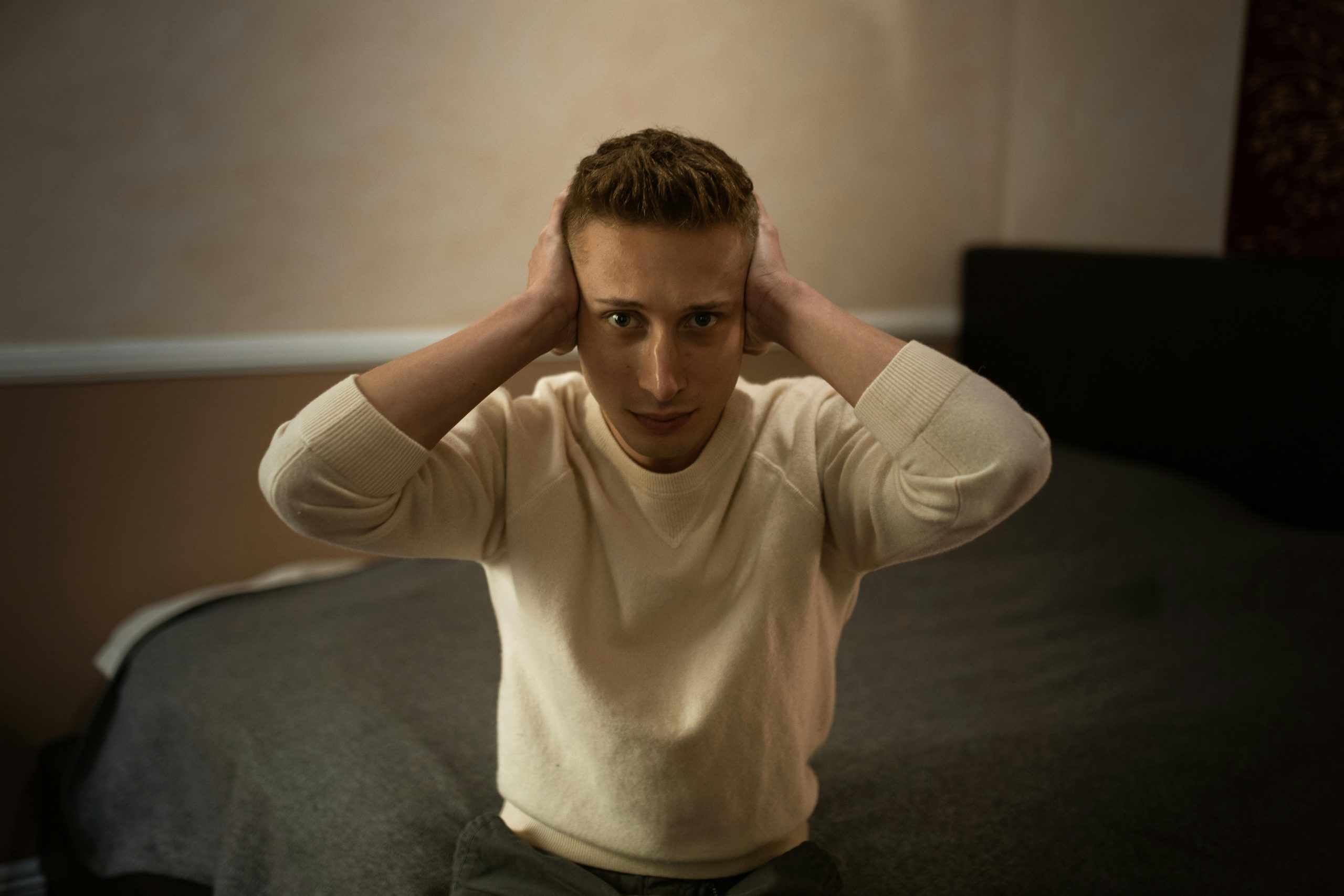 A young man sits on a bed with a distressed expression, covering his ears.