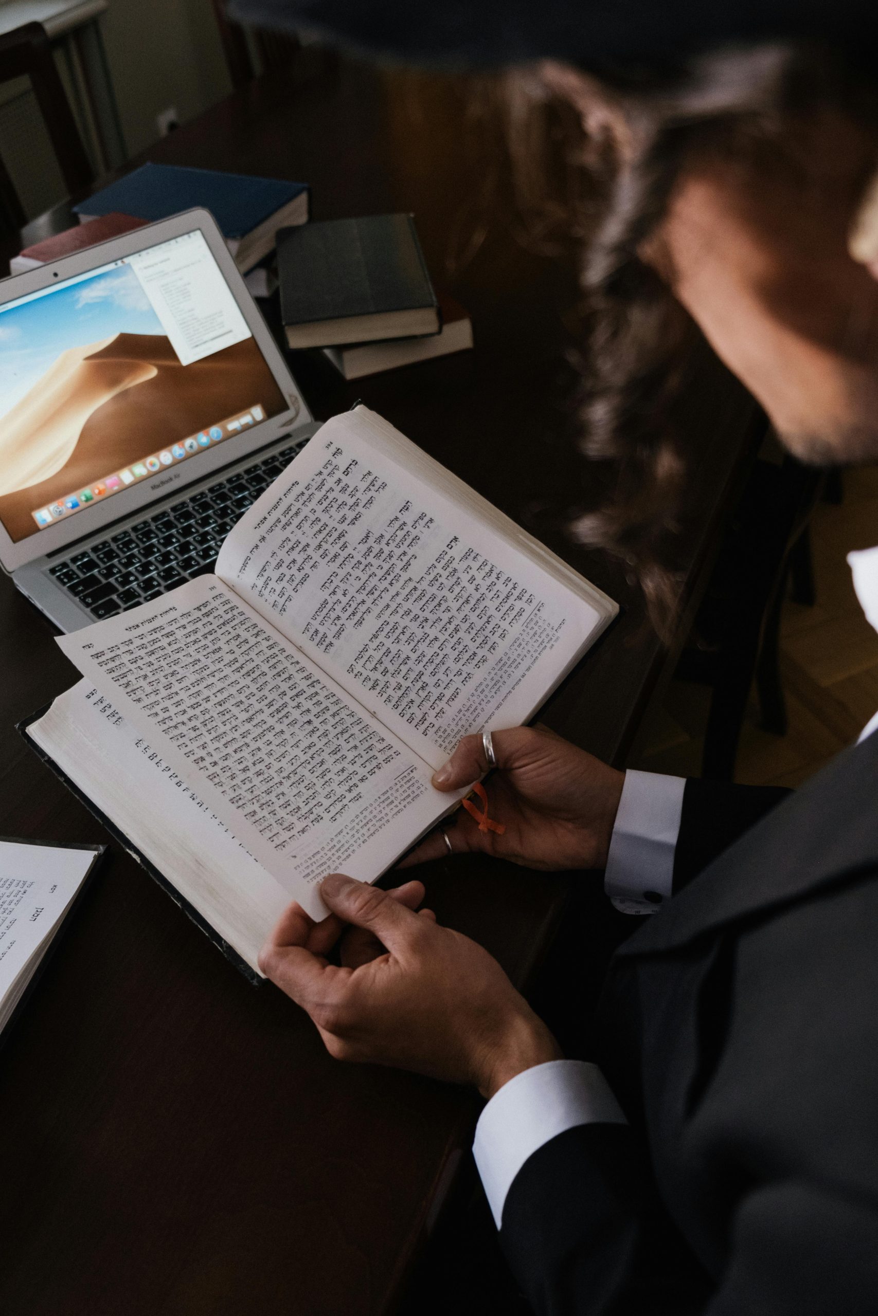 A man in a suit holds a book while working on a laptop at a desk.