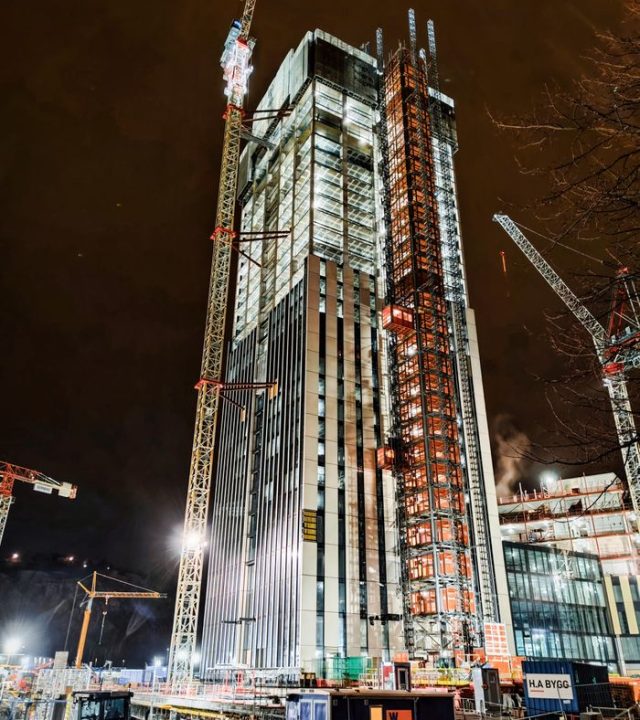 A modern skyscraper under construction at night in Lindholmen, Sweden, illuminated by cranes and lights.
